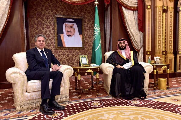Secretary of State Antony J. Blinken and Crown Prince Mohammed bin Salman sitting on chairs in a room with an ornate rug in front of a portrait of King Salman and the green Saudi flag.