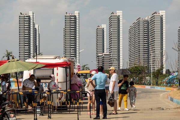 Multistory gray and white apartment towers loom behind people standing near a trailer and a large green umbrella.