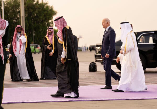 President Biden walking along a purple carpet laid out at an airport. He is surrounded by men in traditional Saudi clothing and head scarves.