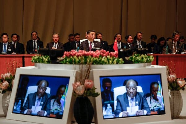 Mr. Xi, in a suit, sits in front of a row of other officials in suits. President Ramaphosa is shown on screens in the foreground.