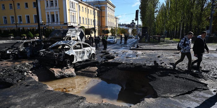 A crater several meters wide in a road in Kyiv on October 10, 2022, after the city was struck by multiple rockets. The scene shows a burnt-out car and people walking.