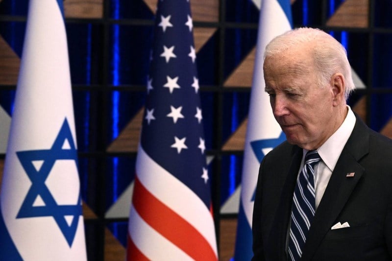 Biden dressed in a dark blue suit walks with his head down past a row of alternating U.S. and Israeli flags.