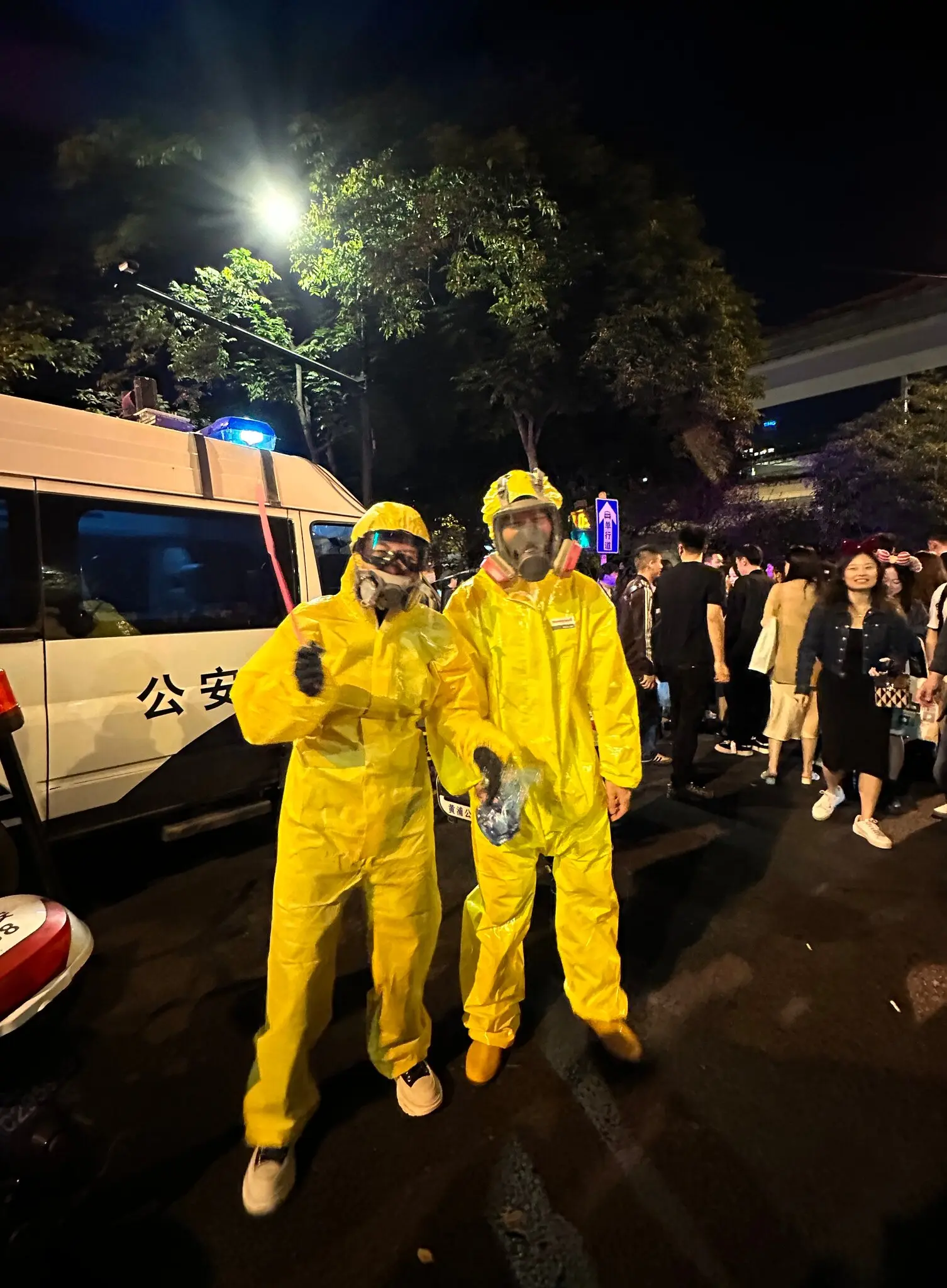 Two people dressed in yellow hazmat suits as characters from the TV show Breaking Bad, standing in front of a crowd and a police car.