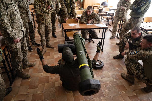 An American volunteer sitting on the ground holding a large Javelin missile surrounded by Ukrainian soldiers who are standing or kneeling.