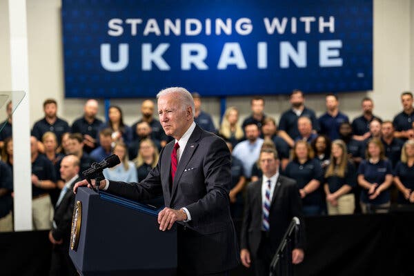 President Biden wearing a suit and standing at a lectern with a crowd of workers behind him and a sign that reads “Standing With Ukraine.”