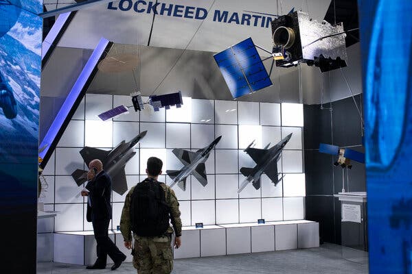 A man in a suit and a man in a military uniform walk past an exposition booth with replica fighter jets and satellites on display and the words “Lockheed Martin.”