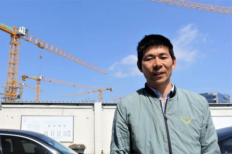 Zhang Xianfeng wears a blue-green jacket and stands in front of a construction site. 