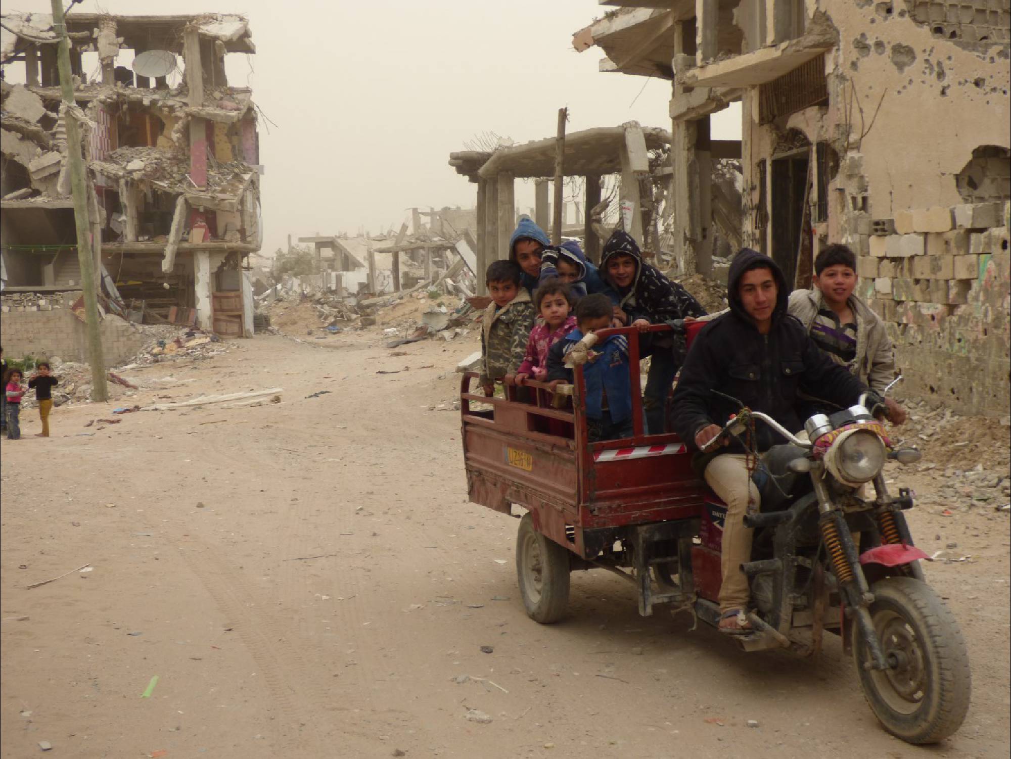 children-riding-on-a-vehicle-with-damaged-buildings-from-hostilities-behind-2000x1502.jpg