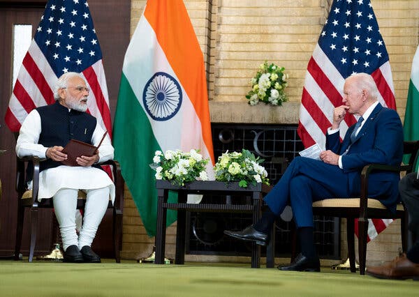 Narendra Modi and President Biden sit opposite each other during a discussion. American and Indian flags hang from poles behind them.
