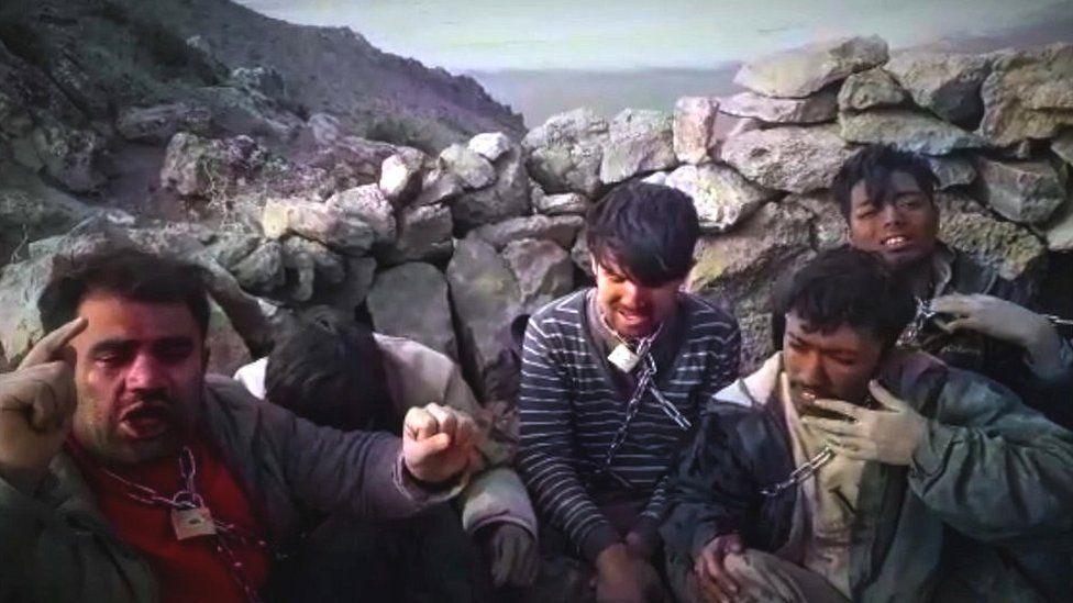 Group of men with chains around their necks sit by a wall on a mountain top