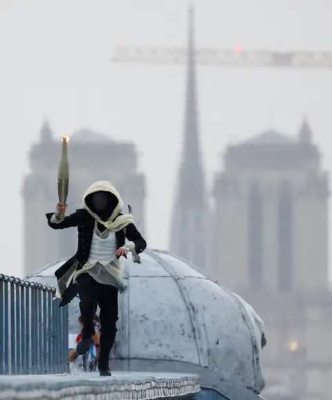 Peter Cziborra/Pool/Getty Images  A  masked torchbearer runs atop the Musee d'Orsay with the Notre-Dame-de-Paris cathedral in the background during the Opening Ceremony of the Olympic Games Paris 2024 on July 26, 2024