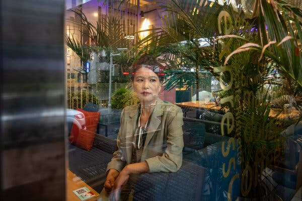 Behind a reflective window, Sun Junli sits on a blue couch inside a restaurant decorated with green foliage. 