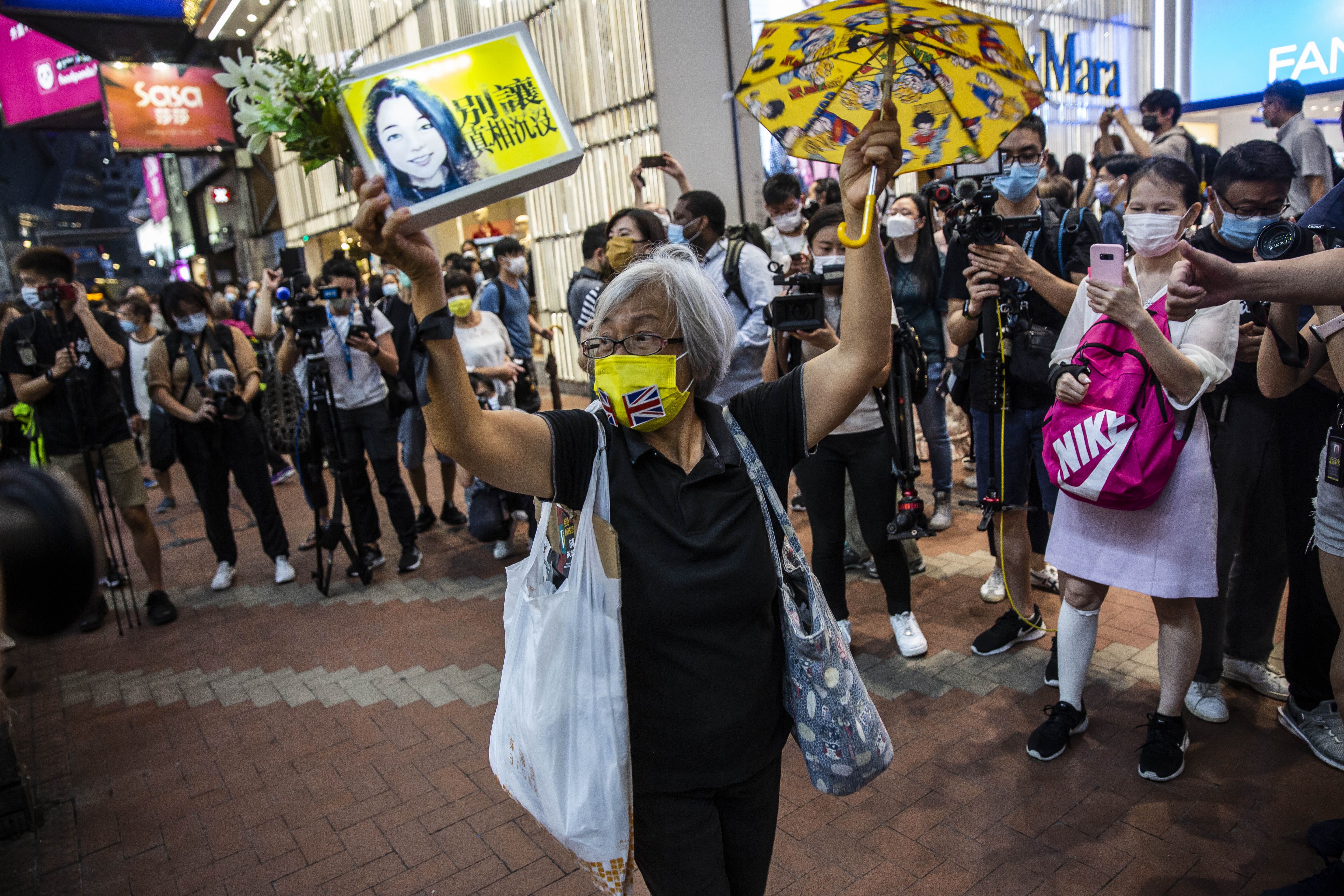 Photo of a masked elderly lady holding a yellow umbrella and sign while surrounded by protesters