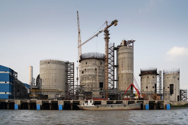 Five cylindrical, concrete structures, along with two construction cranes, tower above a waterfront beside a gray-and-white barge. 