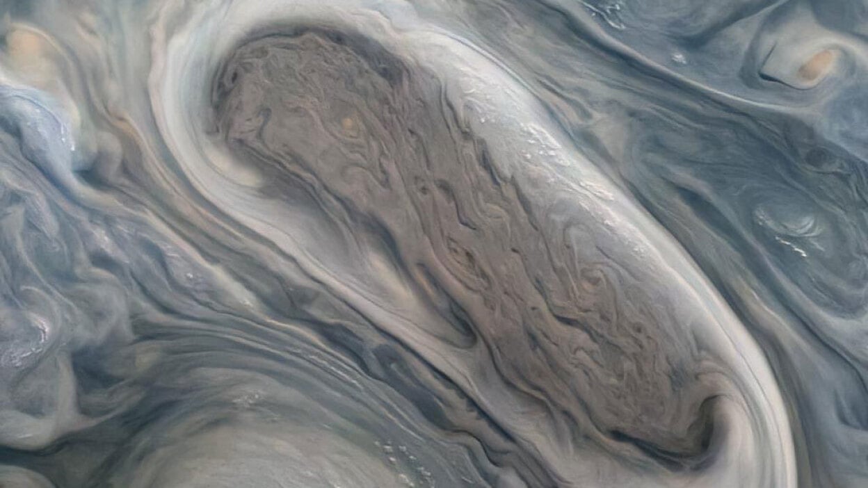 A cropped version of an image captured by NASA's Juno orbiter showing one of Jupiter's massive rotating storms — a swirl of gray-and-blue-tinged clouds — as viewed from orbit.