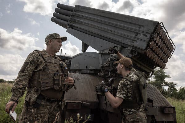 Two Ukrainian soldiers in combat fatigues stand next to artillery weaponry.