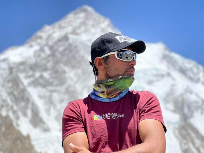 Sirbaz Khan, in t-shirt and sunglasses, poses with K2 in back ground on a sunny day in K2 Base Camp