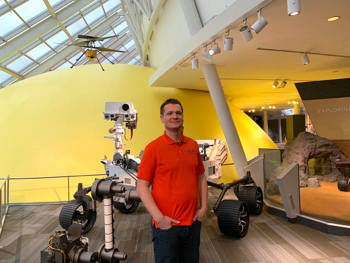 havard grip man in red shirt stands with hands in pockets in front of model of perseverance mars rover with mars helicopter hanging above