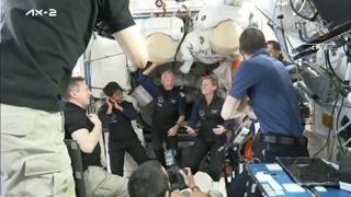 seven smiling people in dark shirts float inside the white-walled international space station.