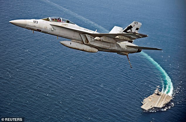 An F/A-18F Super Hornet jet flies over the USS Gerald R. Ford as the Navy aircraft carrier tests its EMALS magnetic launching system, which replaces the steam catapult, in 2017