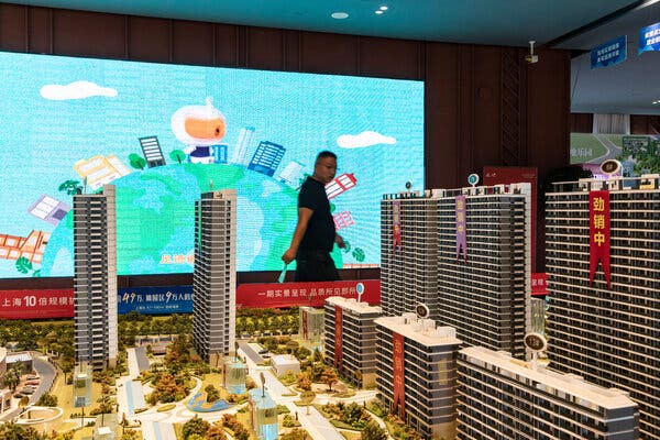 A man walks past a sprawling model of a large apartment-tower complex. Behind him is a bright-blue video screen displaying colorful, cartoon-like images. 