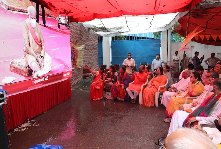 A roomful of seated spectators watches a big screen as India prime minister, Narendra Modi, lays the foundation stone for a new Hindu temple in Ayodhya, August 2021.