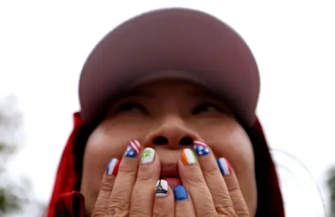  Steph Chambers/Getty Images  A woman wearing a cap covers her mouth with her hands. Her nails are painted with  flags and symbols
