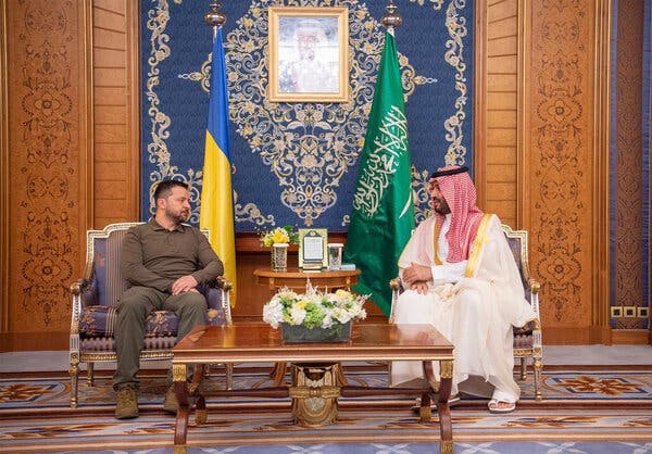 A man in earth-toned clothing sitting in a chair with a Ukrainian flag behind him, alongside a man in white with the Saudi flag behind him.