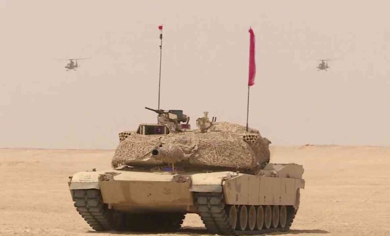 لقطة لإحدى دبابات القتال الرئيسية من طراز M1A1 أبرامز يحلق أعلاها مروحيتي هجوم خفيف من طراز جازيل