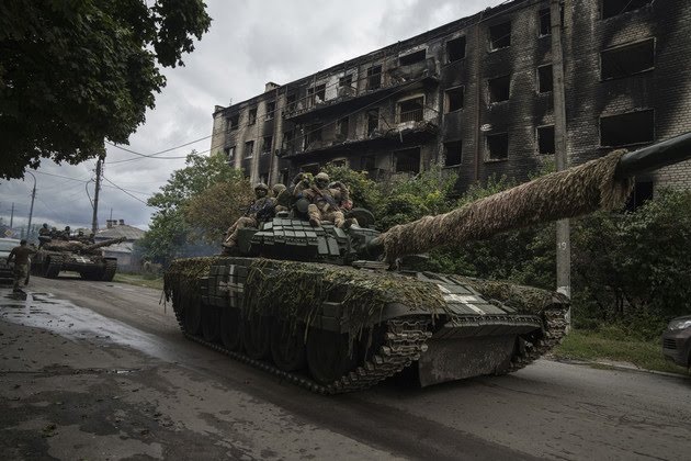 Ukrainian servicemen drive atop a tank that is covered in green moss and dirt. 