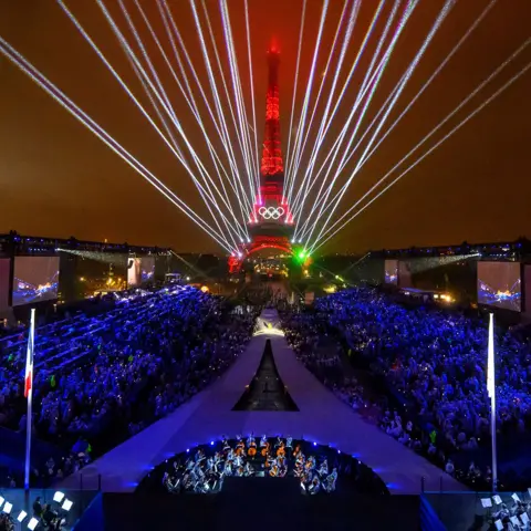François-Xavier Marit/AFP Lights and lasers fill the night sky over the Trocadero venue, with the Eiffel Tower looming in the background and audience members in the foreground, during the opening ceremony of the Paris 2024 Olympic Games