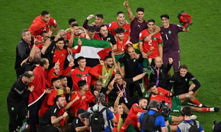 A group of players in red shirts gather for a photograph on the pitch while gesturing in celebration. Several players hold up the Palestine flag in the centre of the group