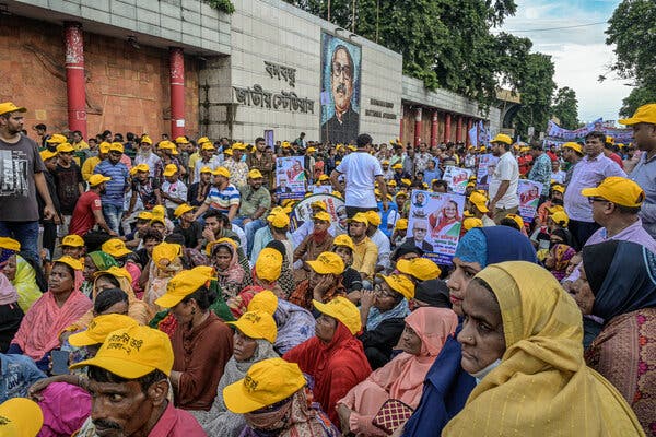 Many people wear the same yellow caps at a political rally. Some are sitting on the street, while others stand. A portrait is painted on a wall in the background. 