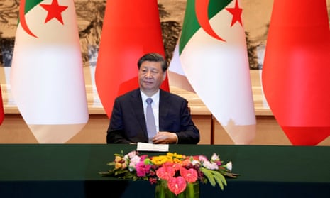 Xi Jinping sits at a black table with flowers on it, and Algerian and Chinese flags hanging behind.