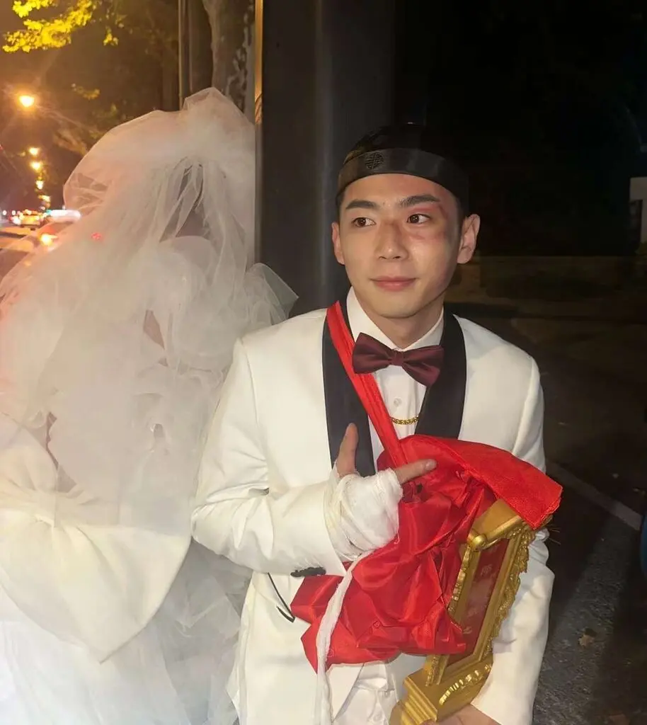 A man dressed up as a groom in tuxedo next to someone dressed up as a bride. He’s wearing a traditional Chinese hat and wedding sash. 