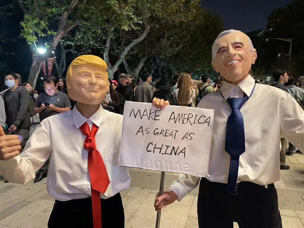 Two people, one wearing a Donald Trump mask another a Joe Biden mask, holding a sign says “Make America as great as China.”