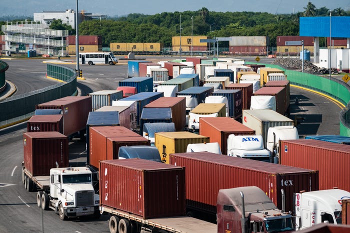 Trucks carrying shipping containers lining up at a checkpoint near on a wide, five-lane road.