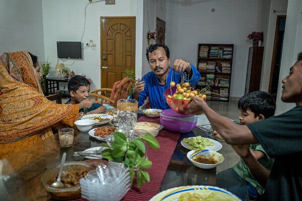 Five people from a family sit at a table during breakfast. One person is handing a bowl filled with food to the father, who is sitting at the end of the table.